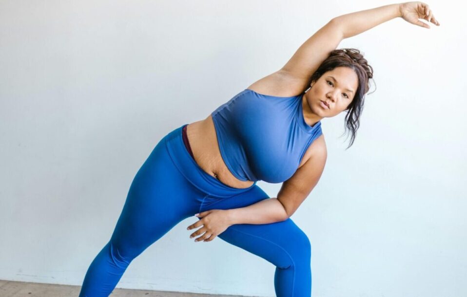 A confident plus size woman in blue activewear performing a stretching exercise indoors, promoting fitness and body positivity.