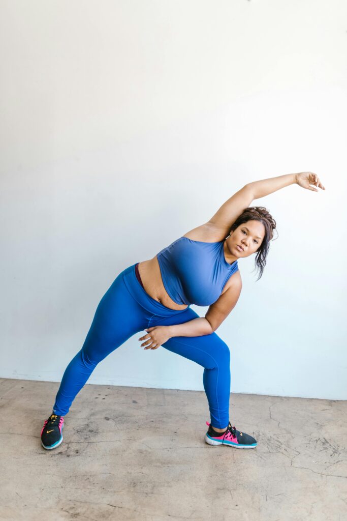 A confident plus size woman in blue activewear performing a stretching exercise indoors, promoting fitness and body positivity.