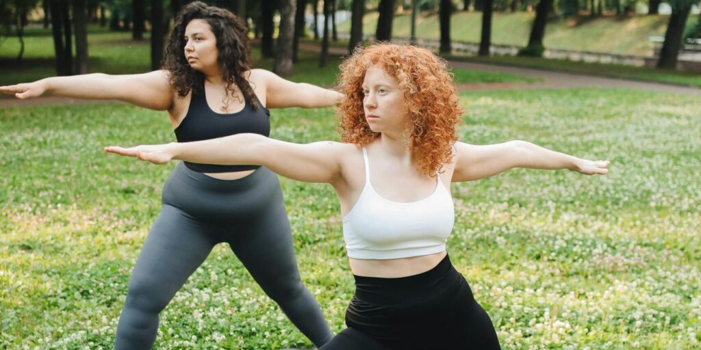 Two women doing yoga poses on mats in a verdant park.